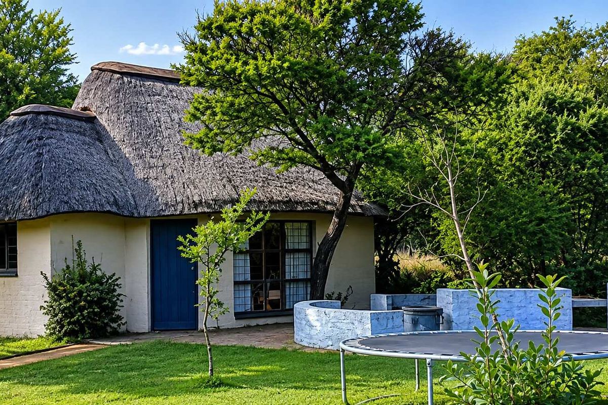 A charming thatched-roof cottage set in a lush garden, featuring a paved patio area with a built-in outdoor braai facility.