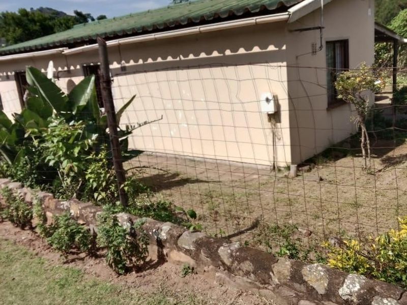 Rear view of a single-story house featuring a corrugated metal roof, stone retaining wall, and wire mesh fencing in a garden setting.
