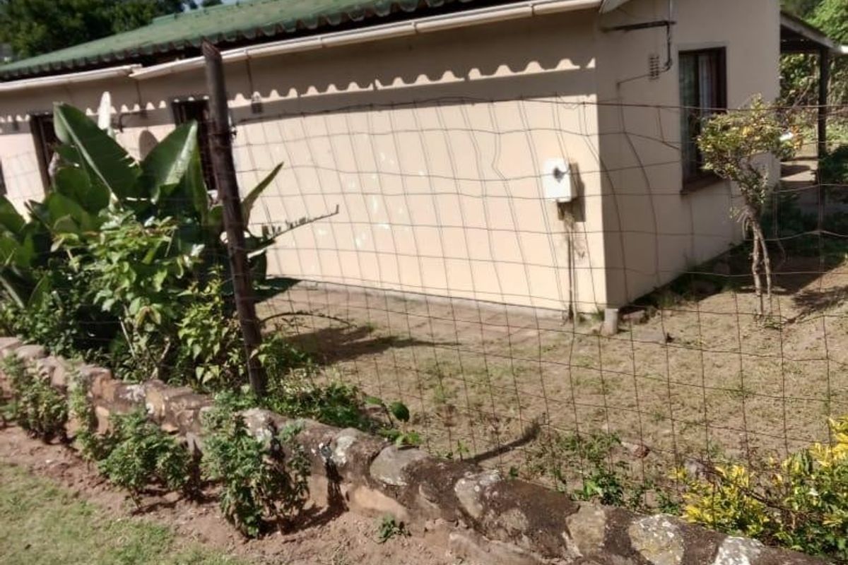 Rear view of a single-story house featuring a corrugated metal roof, stone retaining wall, and wire mesh fencing in a garden setting.