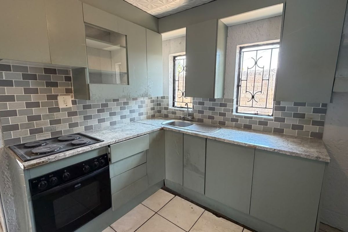 A compact kitchen featuring sage green cabinetry, a built-in oven and hob, tiled backsplash, and security-barred windows, offering a functional layout ready for updates.