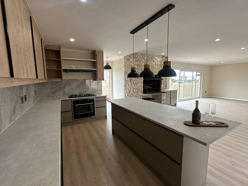 Modern open-plan kitchen featuring stone-look countertops, built-in appliances, and a striking stone-clad fireplace, flowing seamlessly into a spacious living area with sliding door access.