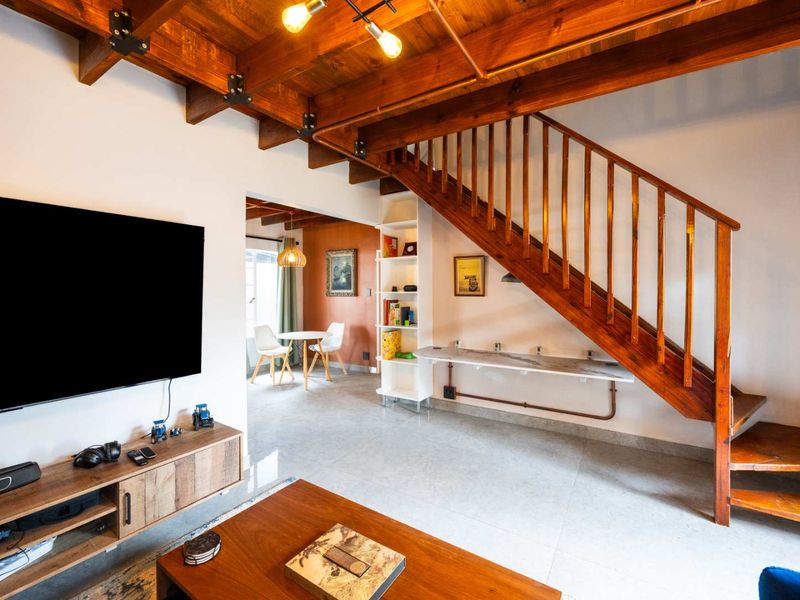 Open-plan living area featuring exposed timber ceiling beams, a wooden staircase, built-in shelving, and a wall-mounted desk, leading to a dining space.
