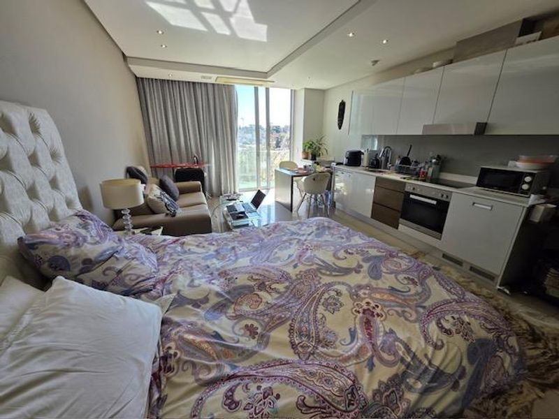 A modern open-plan studio apartment featuring a compact kitchen with built-in cabinetry and integrated appliances, flowing into a combined living and sleeping area.