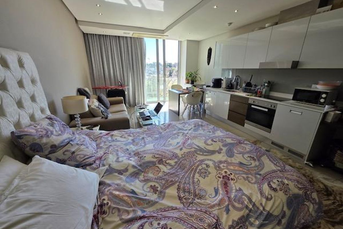 A modern open-plan studio apartment featuring a compact kitchen with built-in cabinetry and integrated appliances, flowing into a combined living and sleeping area.