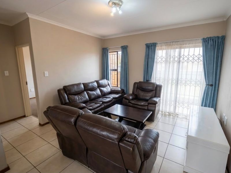 A well-maintained living room featuring neutral wall tones, ceramic floor tiles, and a sliding glass door with security gate providing access to the exterior.