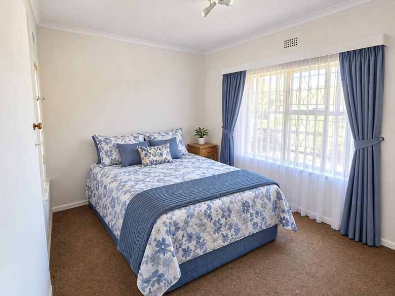 A well-lit bedroom featuring neutral walls, carpeted flooring, and a large window fitted with security bars and curtains.