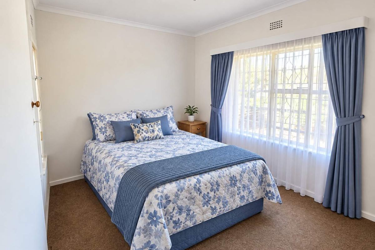 A well-lit bedroom featuring neutral walls, carpeted flooring, and a large window fitted with security bars and curtains.