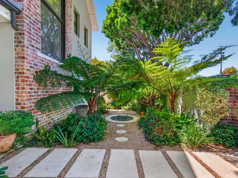 A beautifully landscaped garden area featuring a gravel path, stone pavers, and a decorative metal archway set against a classic brick exterior wall.