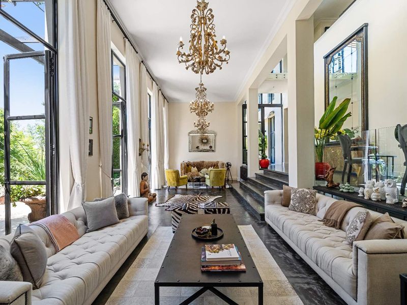 Spacious living room featuring high ceilings, large floor-to-ceiling windows, elegant chandeliers, and a split-level layout with polished dark flooring.