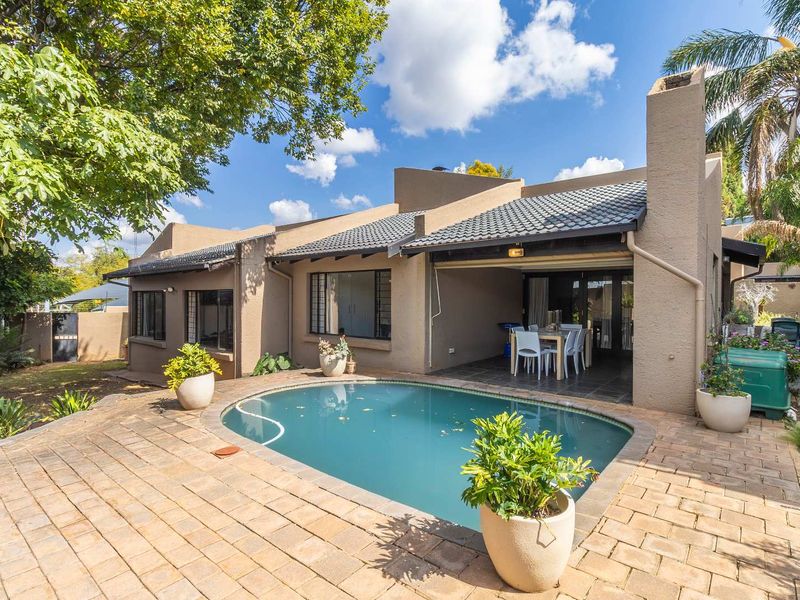 Rear view of the property featuring a covered patio, paved pool deck, and a swimming pool set within a landscaped garden.