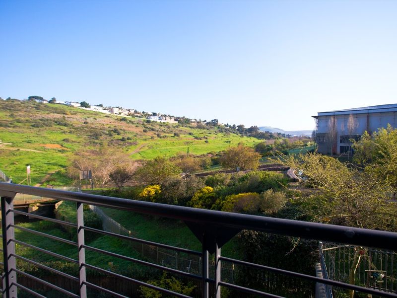 A scenic elevated view overlooking a green hillside, natural landscape, and nearby buildings, captured from a balcony with a metal balustrade.