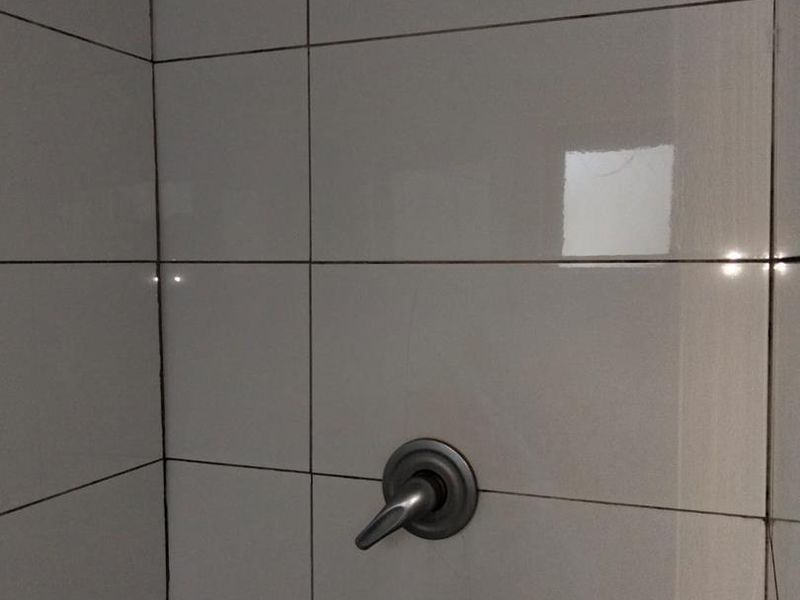 A close-up view of a shower enclosure featuring white ceramic wall tiles, a shower head, and a mixer tap.