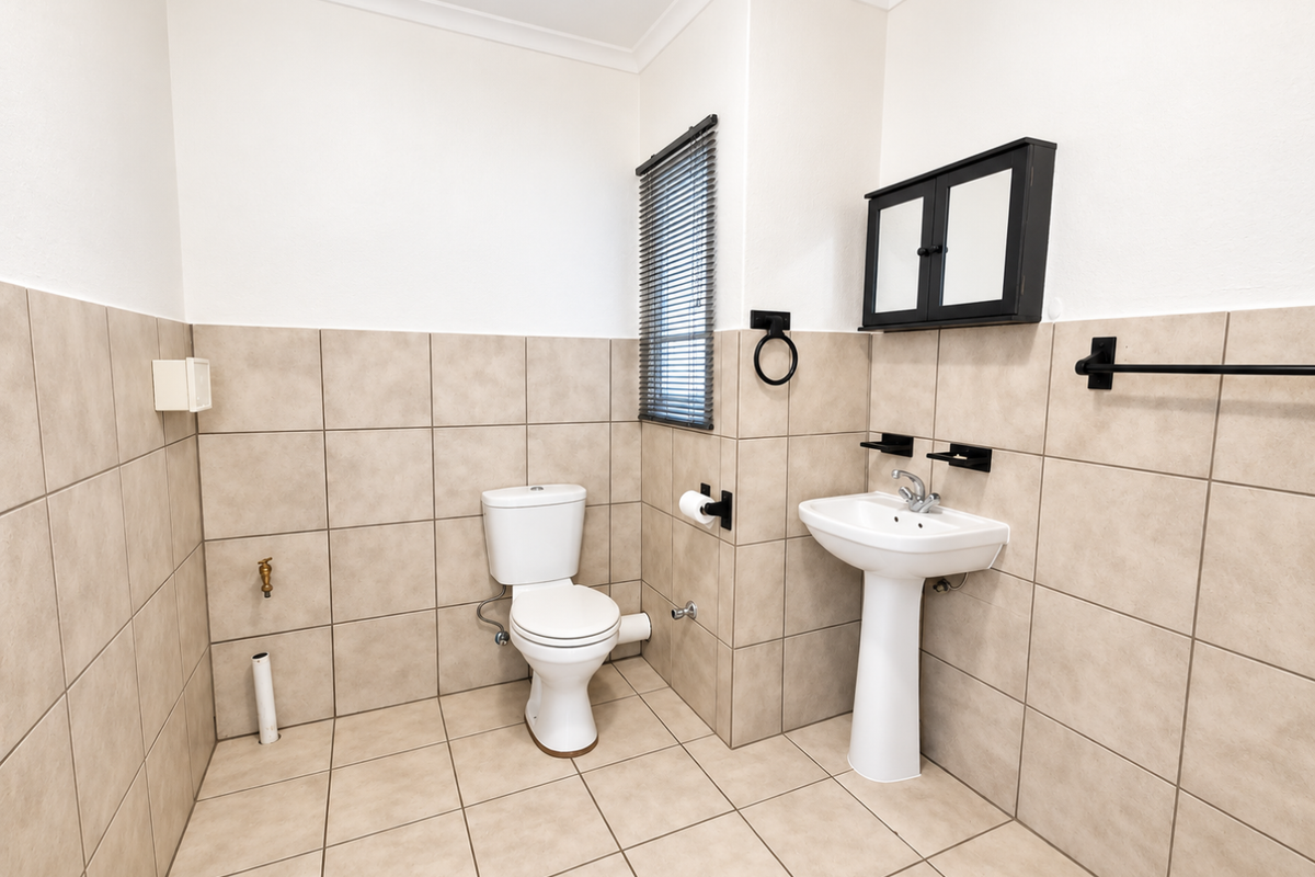 A clean, functional bathroom featuring a pedestal basin, toilet, and neutral wall and floor tiling with modern black fixtures.