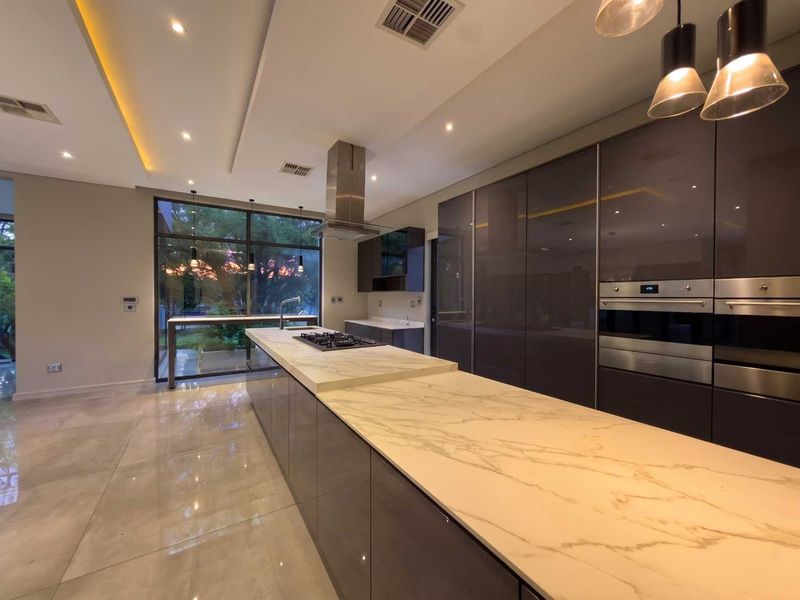 Modern, high-end kitchen featuring a large stone-topped island, integrated appliances, sleek dark cabinetry, and expansive windows overlooking the garden.
