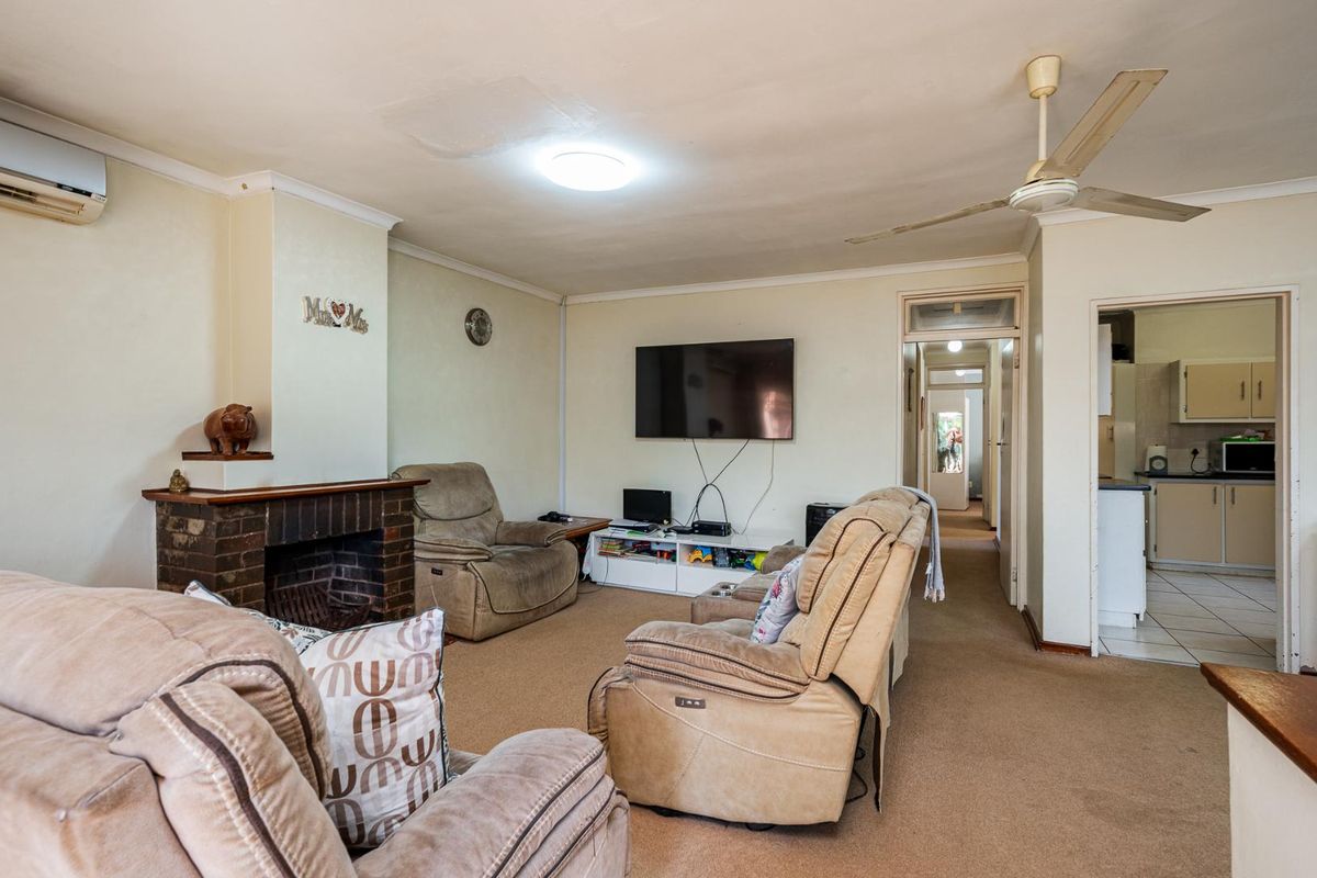 A living room featuring a brick fireplace, ceiling fan, and wall-mounted air conditioning unit, with a view leading into the kitchen area.