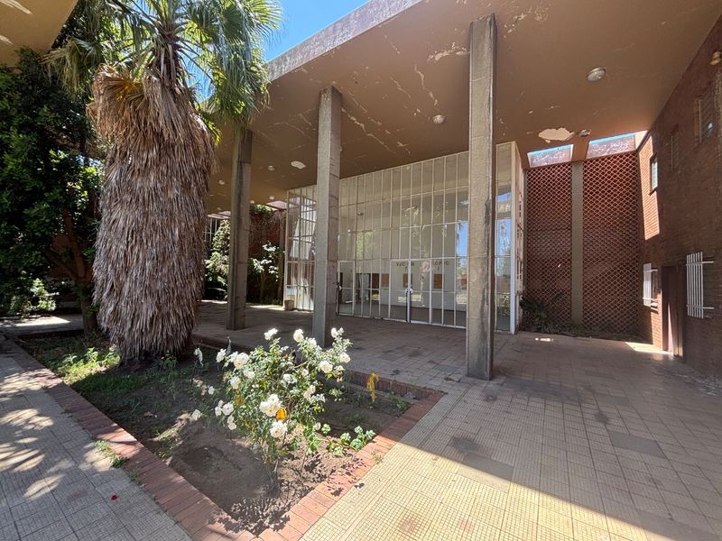 Front entrance of the office building featuring a large glass facade, concrete pillars, and a covered tiled patio area with decorative brickwork.