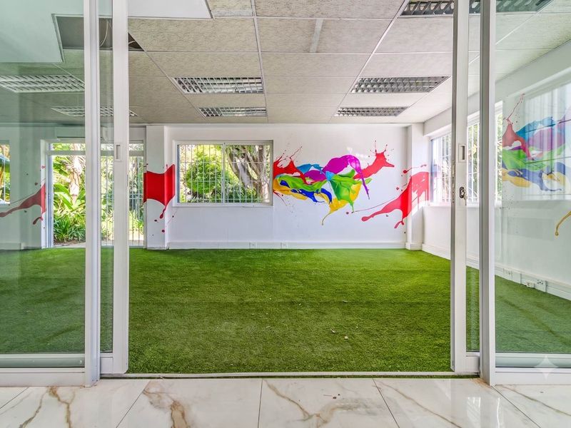 Bright office space featuring artificial turf flooring, suspended ceiling with recessed lighting, and sliding glass doors opening to an exterior view.