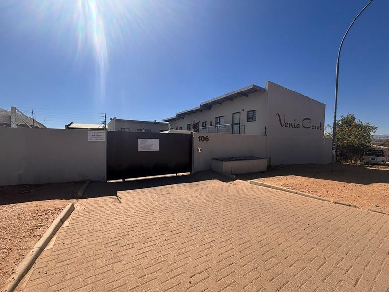Front exterior view of Venia Court apartment complex featuring a secure gated entrance, paved driveway, and perimeter wall with electric fencing.
