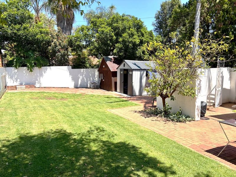 A well-maintained backyard featuring a lush green lawn, brick paved pathways, and a functional storage shed enclosed by a perimeter wall.