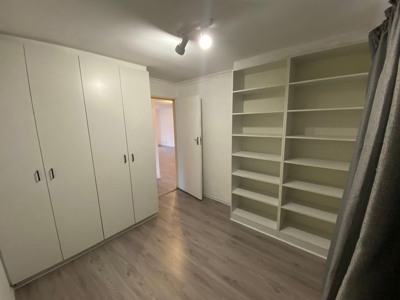 A clean, empty bedroom featuring extensive built-in cupboards, integrated shelving units, and modern laminate flooring, offering a versatile space for storage or study.