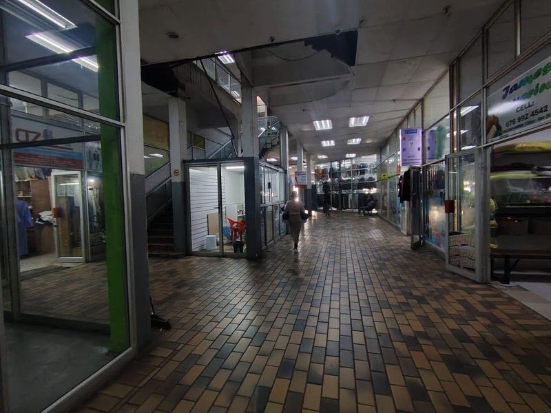 Interior hallway of a retail arcade featuring brick flooring, glass storefronts, and access to upper levels via a staircase.