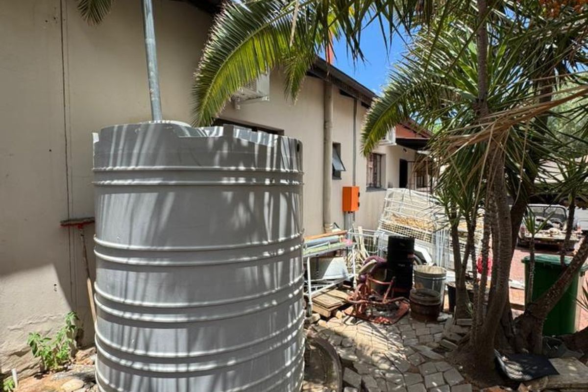 Rear exterior area featuring a large grey water storage tank, paved pathways, and established palm trees alongside the building structure.