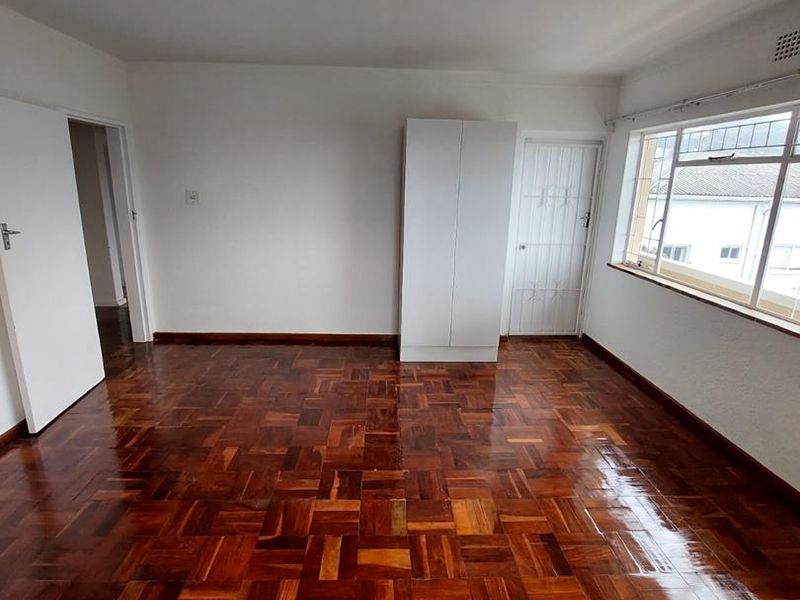 A bright, empty bedroom featuring polished parquet flooring, a built-in wardrobe, and a security gate on the door.