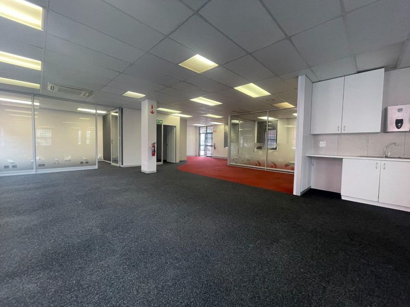 Spacious open-plan office area featuring glass-partitioned meeting rooms, a built-in kitchenette, suspended ceilings, and carpeted flooring throughout.