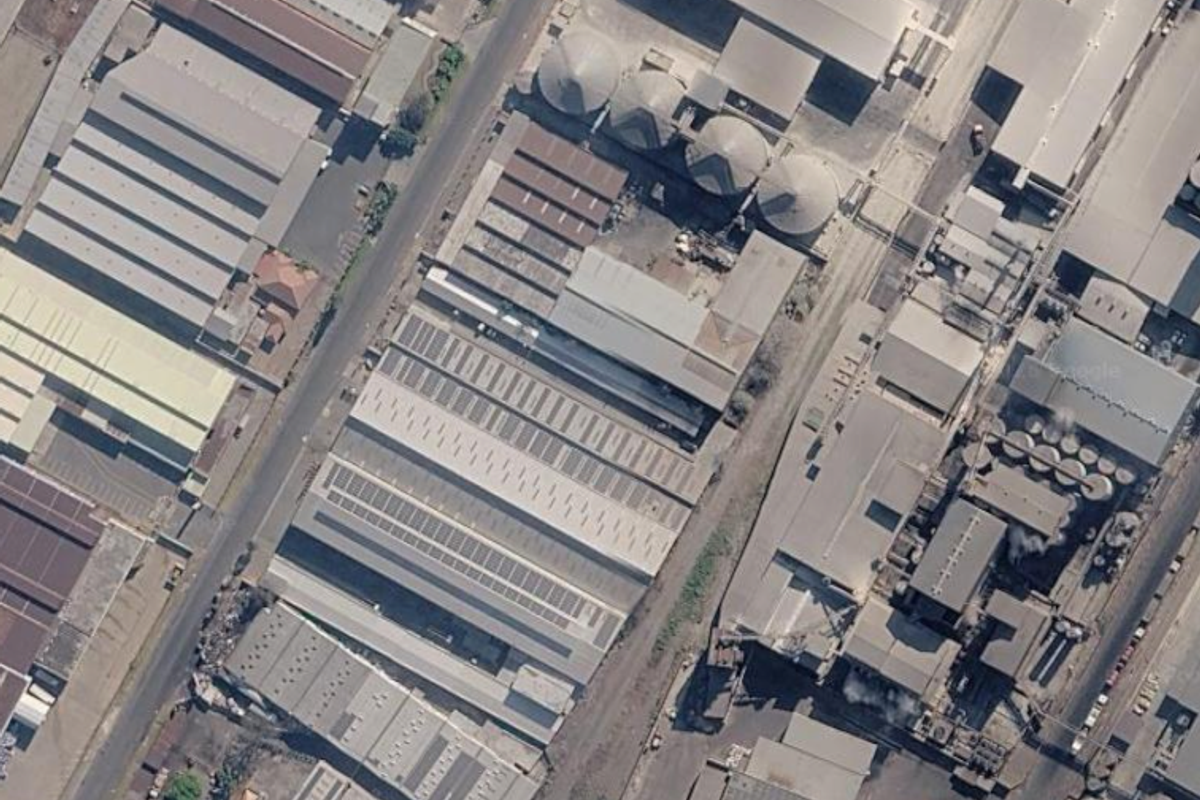 Aerial view of a large industrial facility featuring multiple warehouse structures, silo storage units, and extensive solar panel installations on the roof.