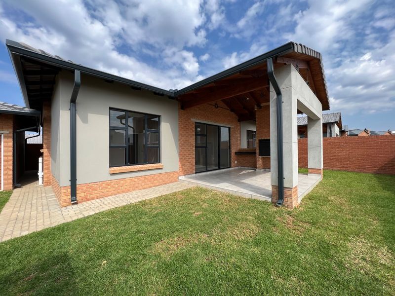 A well-maintained rear exterior view of a townhouse featuring a covered patio with a built-in braai, brick and plaster facade, and a landscaped lawn.