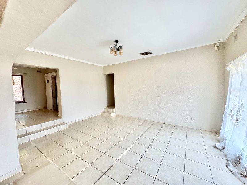 An empty, open-plan living area featuring neutral ceramic floor tiles, coved ceilings, and a stepped transition to an adjacent hallway.