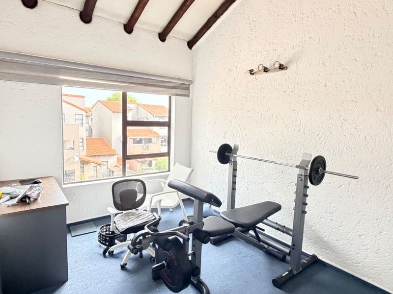 A multi-purpose room featuring exposed timber ceiling beams, textured walls, and a large window with a roller blind, currently utilized as a home office.