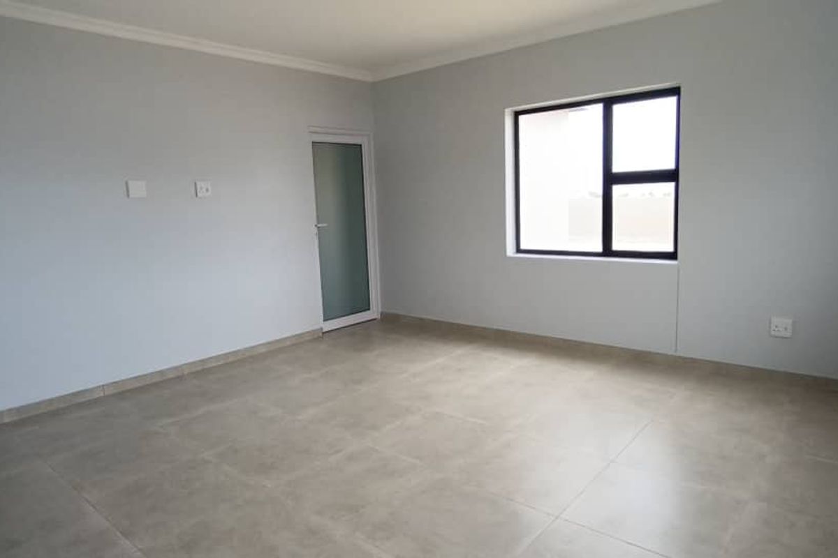 A bright, empty living room featuring neutral tiled flooring, light grey walls, and a modern window with dark frames.