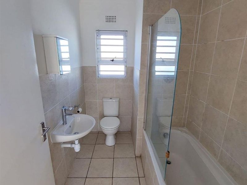 A functional bathroom featuring a wall-mounted basin, toilet, and a bathtub with a glass shower screen, finished with neutral wall and floor tiles.