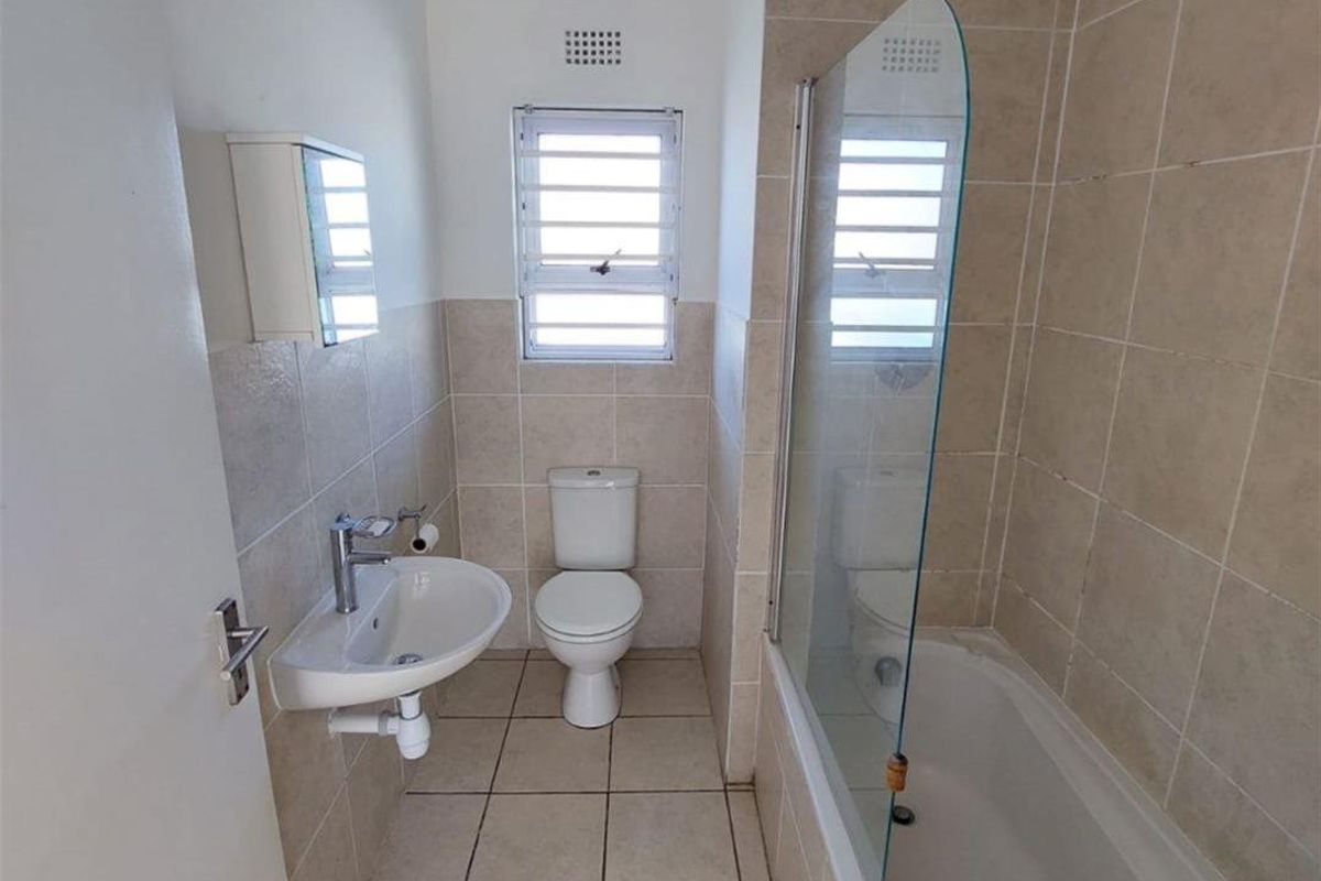 A functional bathroom featuring a wall-mounted basin, toilet, and a bathtub with a glass shower screen, finished with neutral wall and floor tiles.