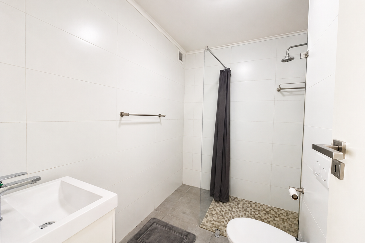 A clean, modern bathroom featuring white wall tiles, a glass shower screen, a wall-mounted basin, and a toilet with neutral floor tiling.