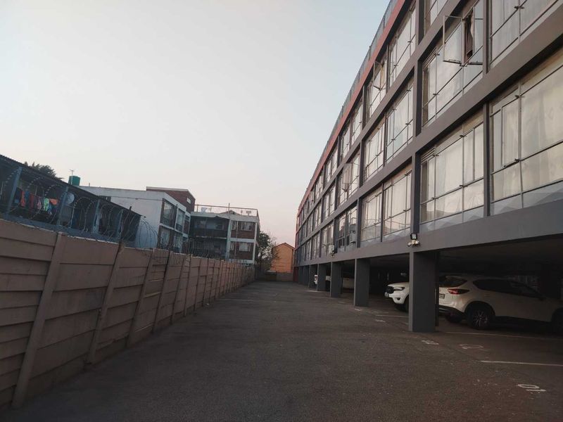 Exterior view of an apartment complex featuring a paved parking area, covered parking bays, and a concrete boundary wall with security fencing.