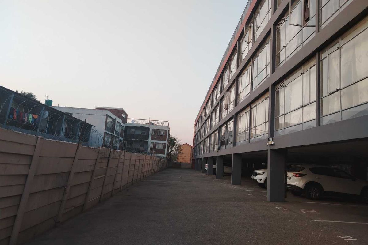 Exterior view of an apartment complex featuring a paved parking area, covered parking bays, and a concrete boundary wall with security fencing.
