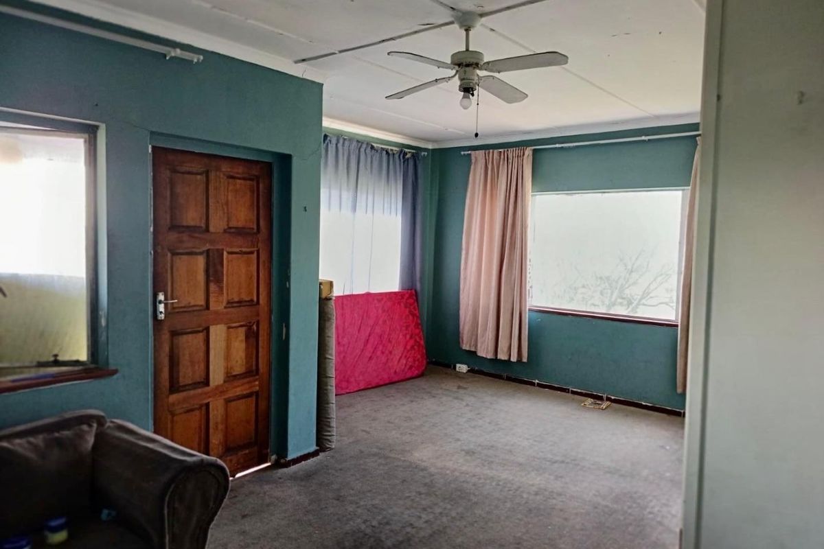 A spacious living room featuring a ceiling fan, carpeted flooring, and large windows, currently in need of cosmetic updates and repairs.