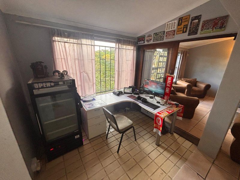 A functional home office space featuring tiled flooring, large windows with security bars, and an open-plan layout connecting to the adjacent living area.