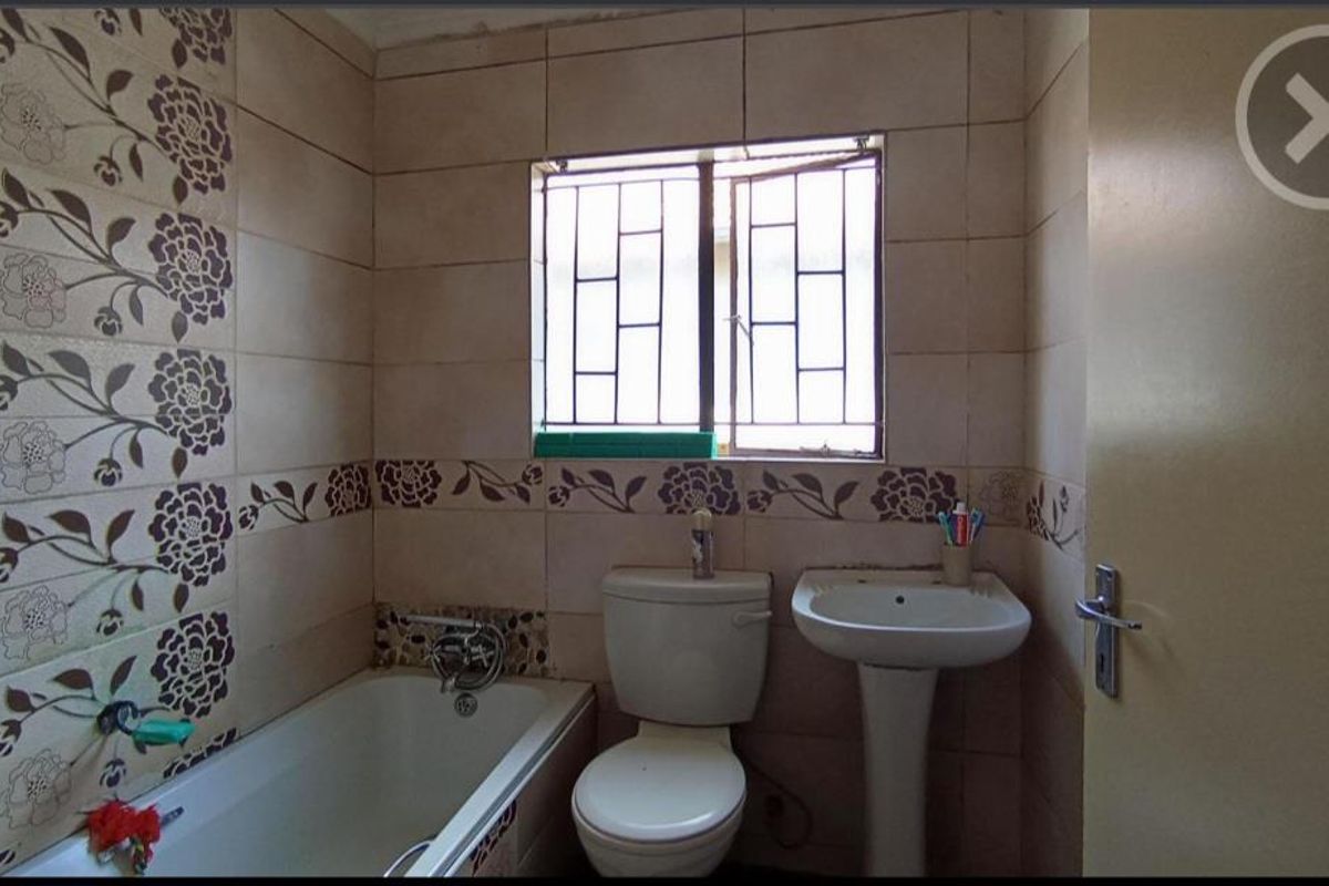 A compact bathroom featuring a standard bathtub, pedestal basin, and toilet, finished with dated floral-patterned wall tiles and a window with security bars.