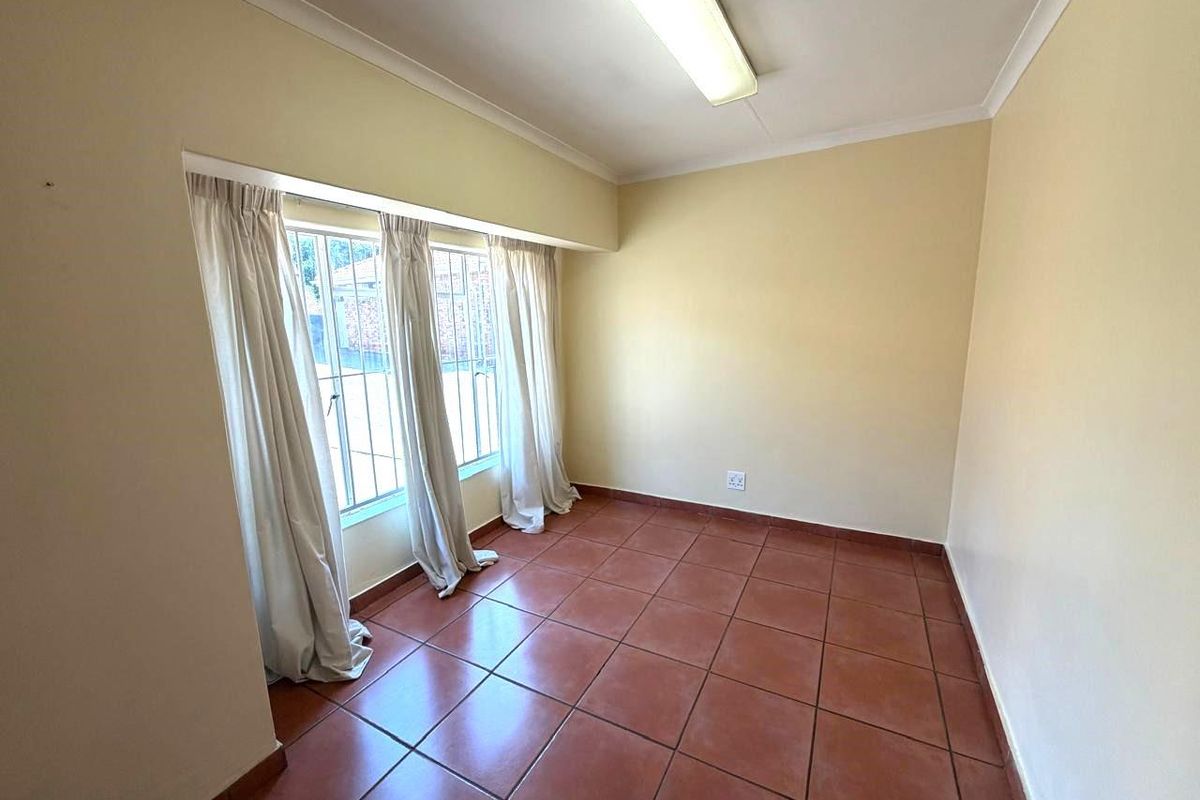 A bright, empty bedroom featuring terracotta floor tiles, large windows with security bars, and a simple ceiling-mounted light fixture.