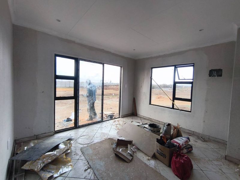 An unfinished living area featuring tiled floors, aluminium sliding doors, and large windows, currently undergoing construction with exposed walls and electrical points.