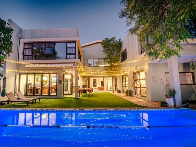 A beautifully illuminated rear exterior view featuring a swimming pool, artificial lawn, and a spacious covered patio area with large glass sliding doors.