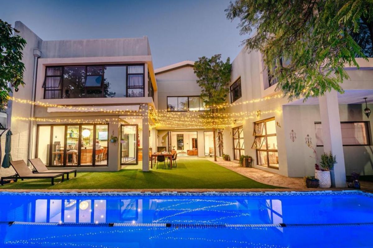 A beautifully illuminated rear exterior view featuring a swimming pool, artificial lawn, and a spacious covered patio area with large glass sliding doors.