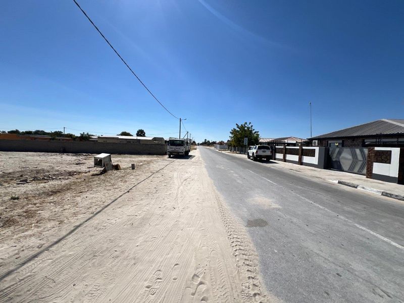 A street view showing the asphalt road, adjacent sandy verge, and residential boundary wall with a modern sliding gate.