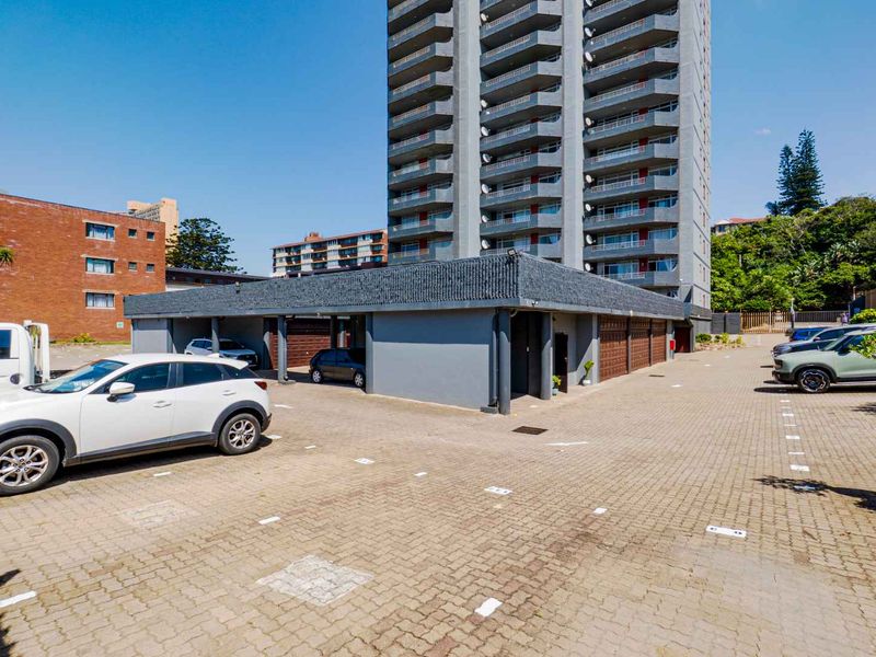 Exterior view of a multi-story apartment complex featuring a paved parking area with covered bays and residential balconies.