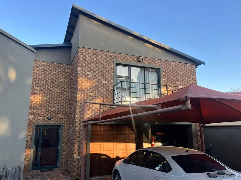 Front elevation of a double-story face brick house featuring a balcony with metal railing, a garage, and a shaded parking area.