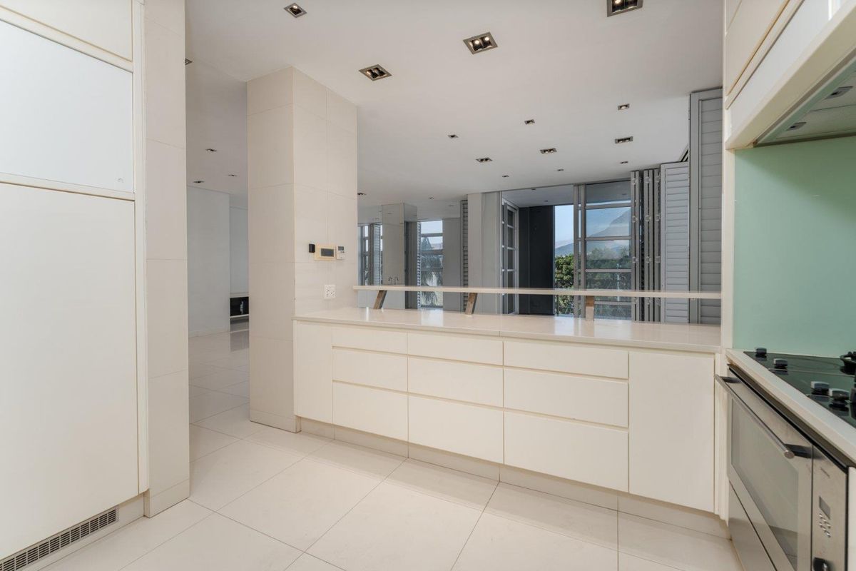 Modern, minimalist kitchen featuring sleek white cabinetry, stone countertops, integrated appliances, and high-quality tiled flooring, opening into a bright, spacious living area.