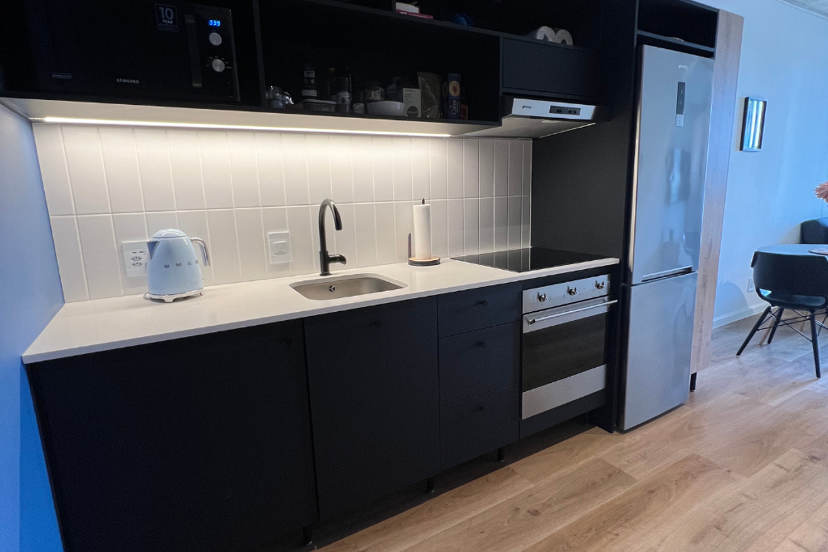 Modern kitchen featuring matte black cabinetry, white tiled backsplash, integrated oven, induction cooktop, and stainless steel refrigerator with light wood-look flooring.